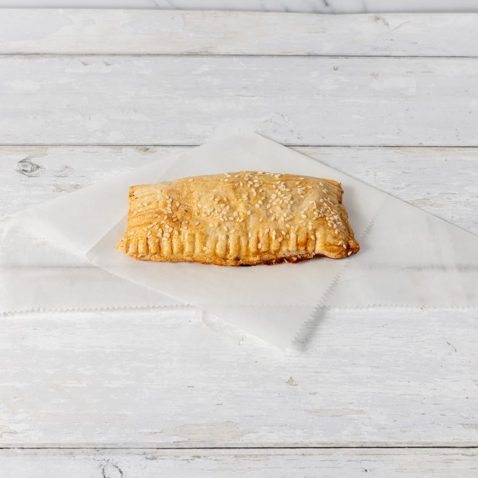 A golden-brown pastry with sesame seeds on a white paper liner, on a distressed white wooden surface.