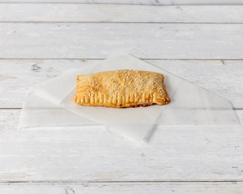 A golden-brown pastry with sesame seeds on a white paper liner, on a distressed white wooden surface.