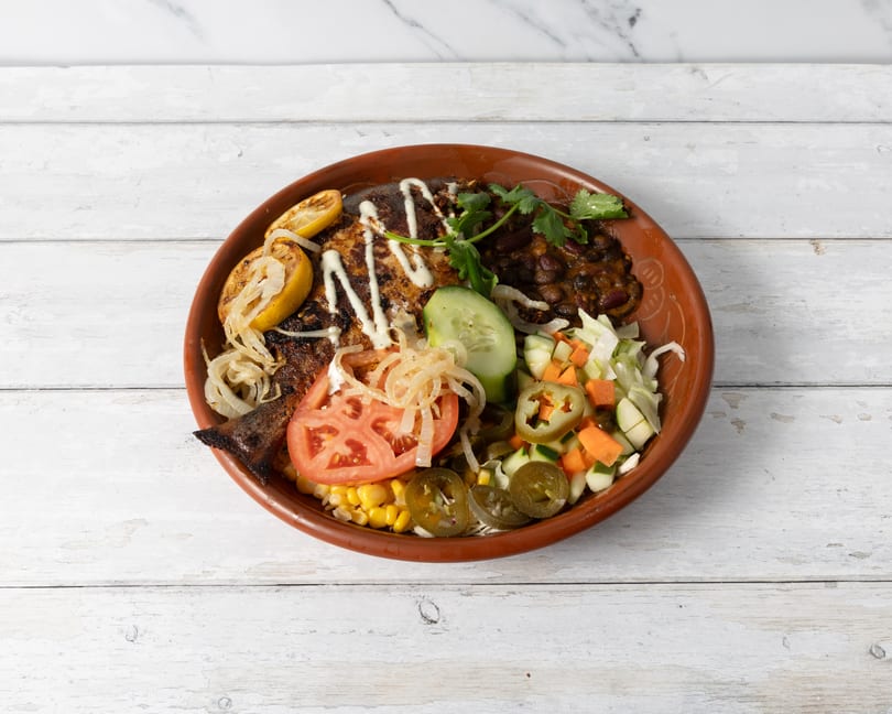 Grilled fish with lemon, beans, corn, tomatoes, cucumbers, and jalapeños in a rustic bowl.