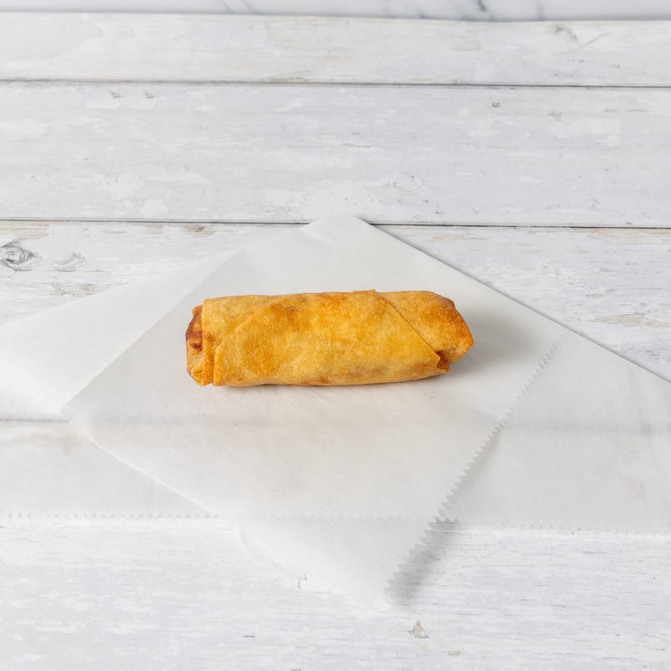 A single golden-brown egg roll rests on a white paper liner against a distressed white wooden background.