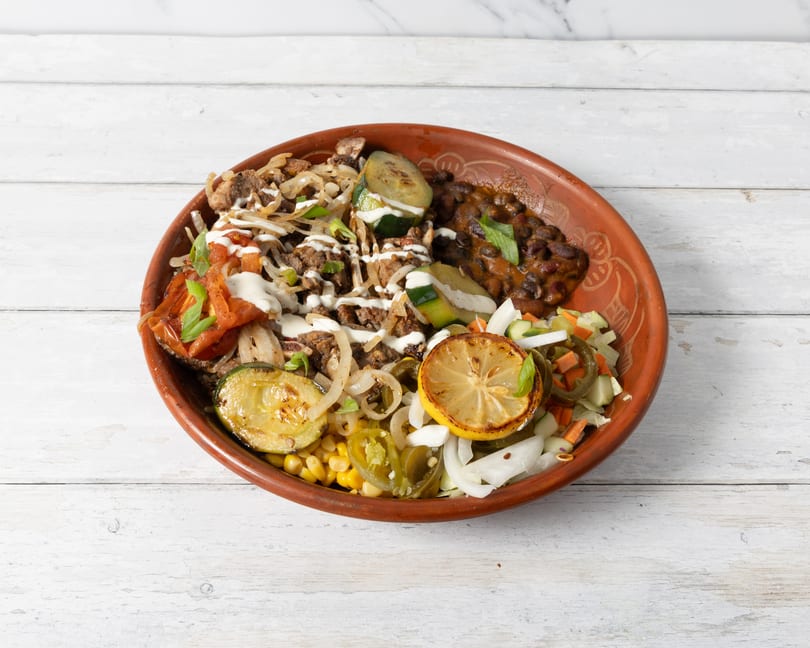 A colorful bowl of food with meat, vegetables, beans, and a grilled lemon on a white wooden table.