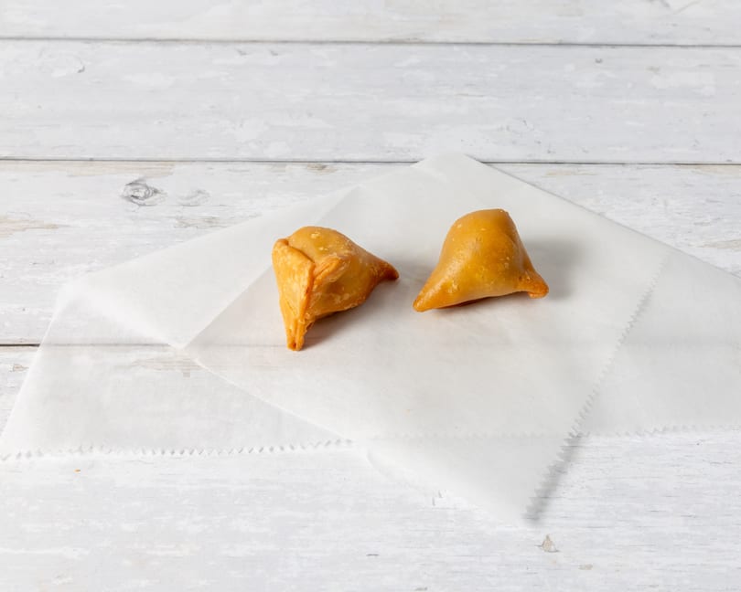 Two golden-brown samosas rest on white parchment paper against a distressed white wooden background.