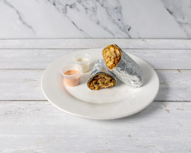 Two foil-wrapped shawarma wraps with chicken and vegetables, served with two dipping sauces on a white plate.
