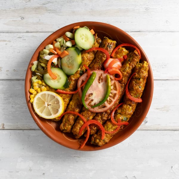 A clay plate of seekh kebabs with a side salad of cucumbers, carrots, corn, and lemon.