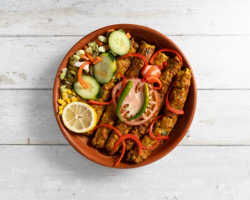 A clay plate of seekh kebabs with a side salad of cucumbers, carrots, corn, and lemon.