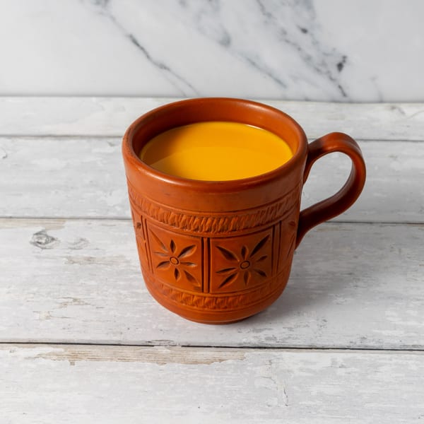 A terracotta mug with carved floral patterns holds a bright yellow beverage on a distressed white wooden surface.