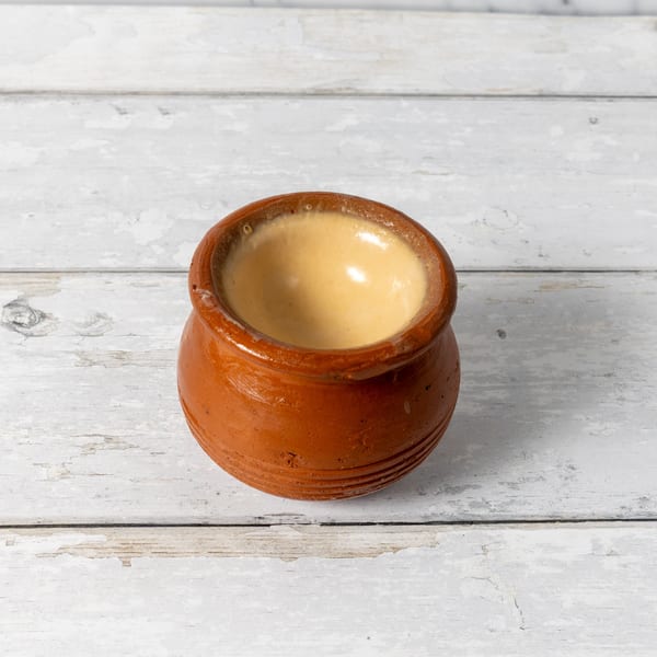 A small, rustic terracotta pot filled with a creamy, light brown dessert on a distressed white wooden surface.
