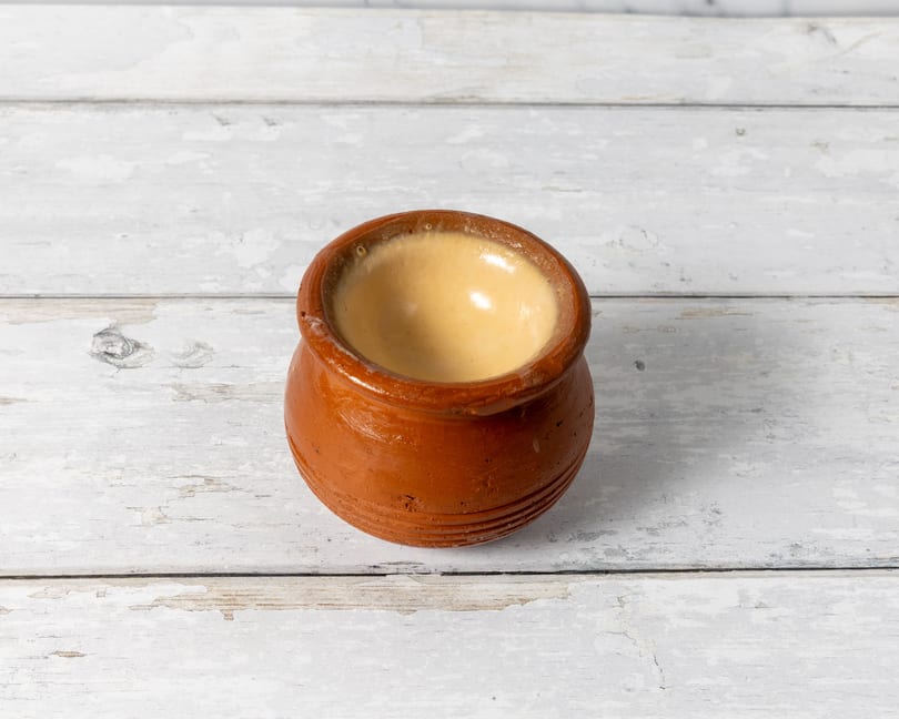 A small, rustic terracotta pot filled with a creamy, light brown dessert on a distressed white wooden surface.