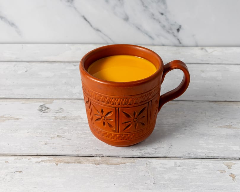 A terracotta mug with carved floral patterns, filled with a bright yellow beverage, on a white wooden surface.
