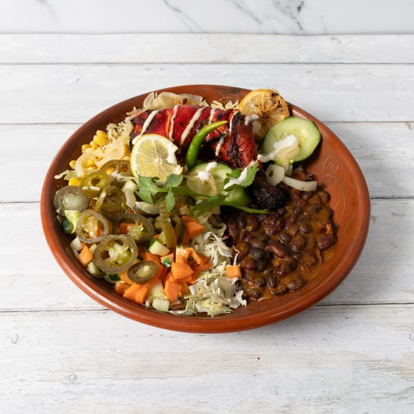 A vibrant platter with red chicken, rice, black beans, corn, pickled jalapeños, and fresh vegetables.