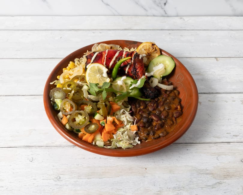 A vibrant platter with red chicken, rice, black beans, corn, pickled jalapeños, and fresh vegetables.