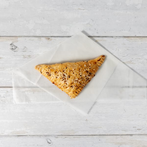 A golden-brown triangular pastry, sprinkled with sesame seeds and spices, rests on white parchment paper.